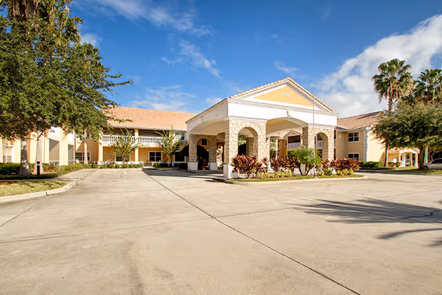 Front entrance of a two-story senior living facility with a covered porte-cochère, landscaped plants, and a wide driveway under a blue sky.