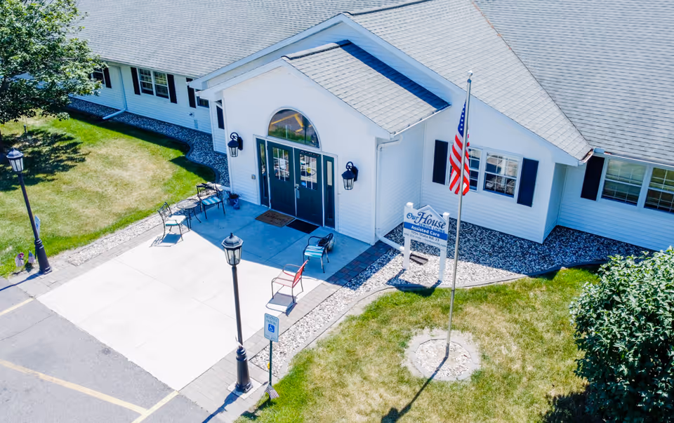 Exterior view of Our House Senior Living - Wisconsin Dells Assisted Care building with white siding, a gray roof, and a front entrance featuring double doors and outdoor seating. There is a flagpole with an American flag and a sign displaying the facility's name. The surrounding area includes a lawn, trees, and a parking lot with a handicapped parking sign.