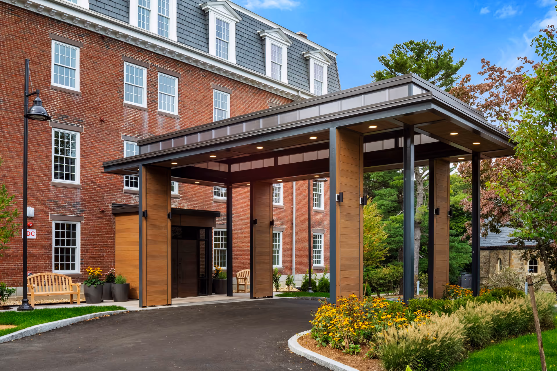 Entrance area of a senior living facility with a covered drop-off zone supported by wooden and metal pillars. The building is made of red brick with multiple white-framed windows. There are benches, potted plants, and landscaped flower beds with yellow flowers and greenery around the entrance. Trees and a clear blue sky are visible in the background.