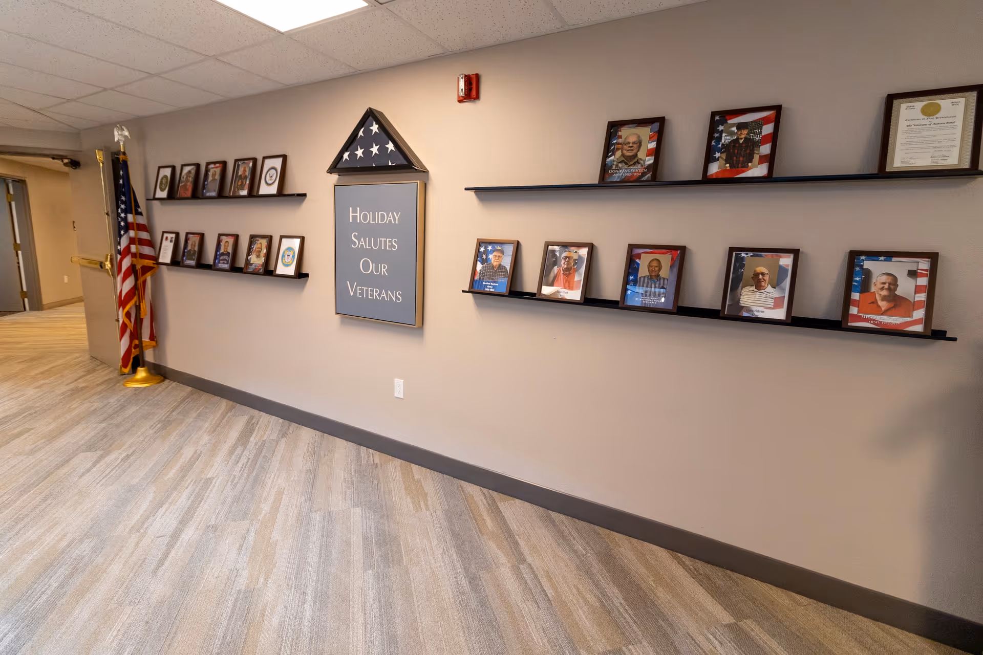 A hallway wall display honoring veterans with framed photos of veterans on black shelves, an American flag in a triangular display case, and a sign that reads 'Holiday Salutes Our Veterans'. The floor is carpeted with a light wood pattern and the walls are painted beige.