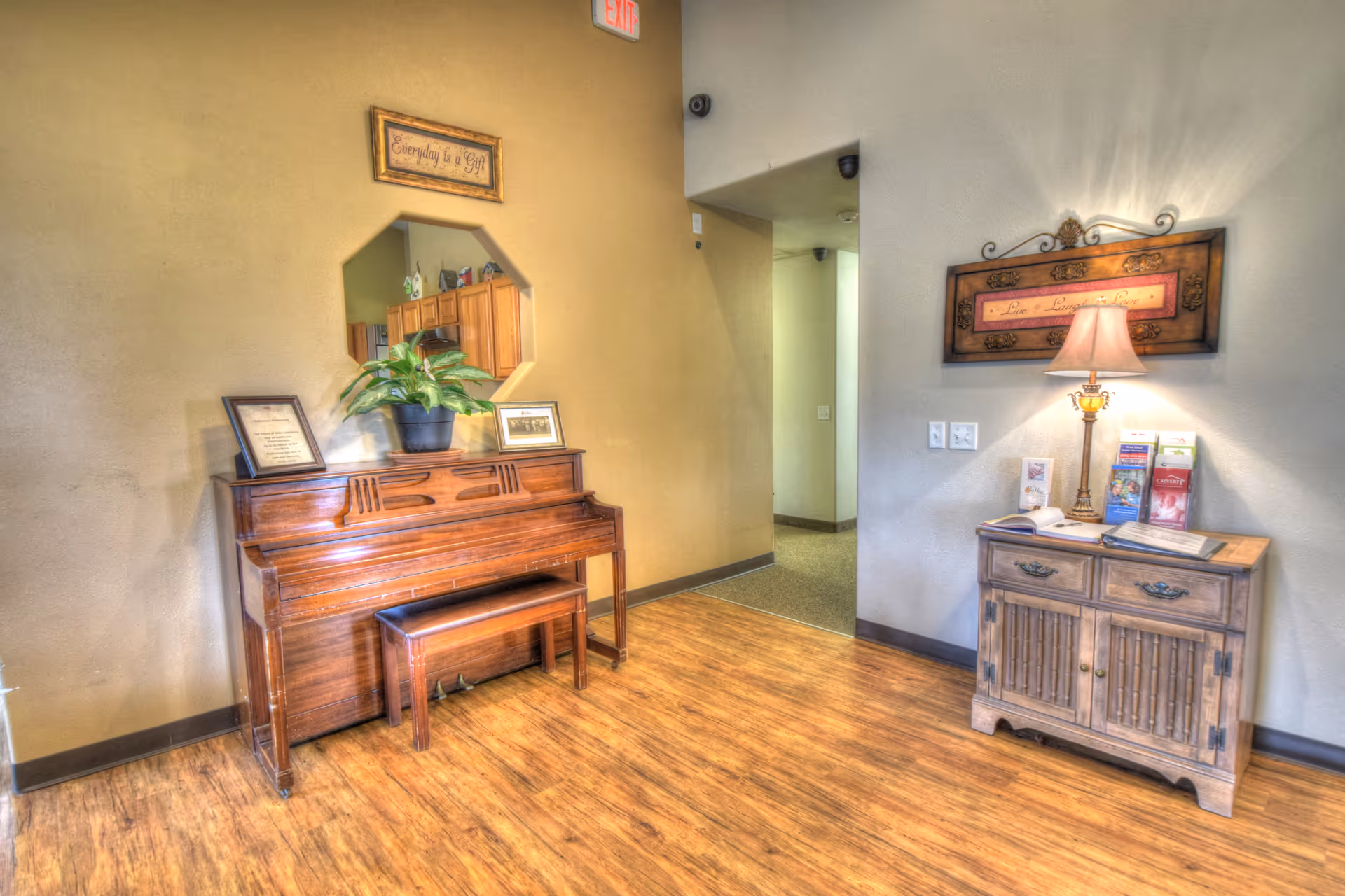 Interior view of a senior living facility hallway corner featuring a wooden upright piano with a bench, a potted plant, and framed pictures on top. Above the piano is a decorative mirror and a framed sign that reads 'Everyday is a Gift.' To the right, there is a wooden cabinet with brochures, a table lamp, and a framed wall decoration with the words 'Live, Laugh, Love.' The floor is wooden, and there is an exit sign visible above a hallway entrance.
