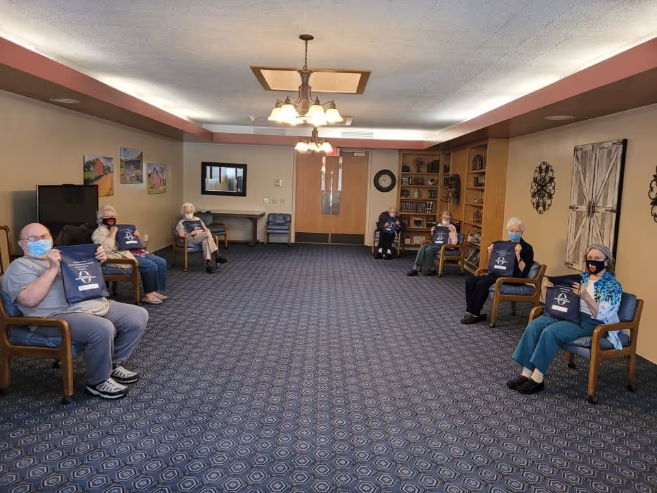 A group of elderly people sitting spaced apart in a large room with blue patterned carpet and beige walls. Each person is wearing a mask and holding a navy blue bag with white text and a logo. The room has wooden chairs, a bookshelf, wall decorations, and overhead lights.