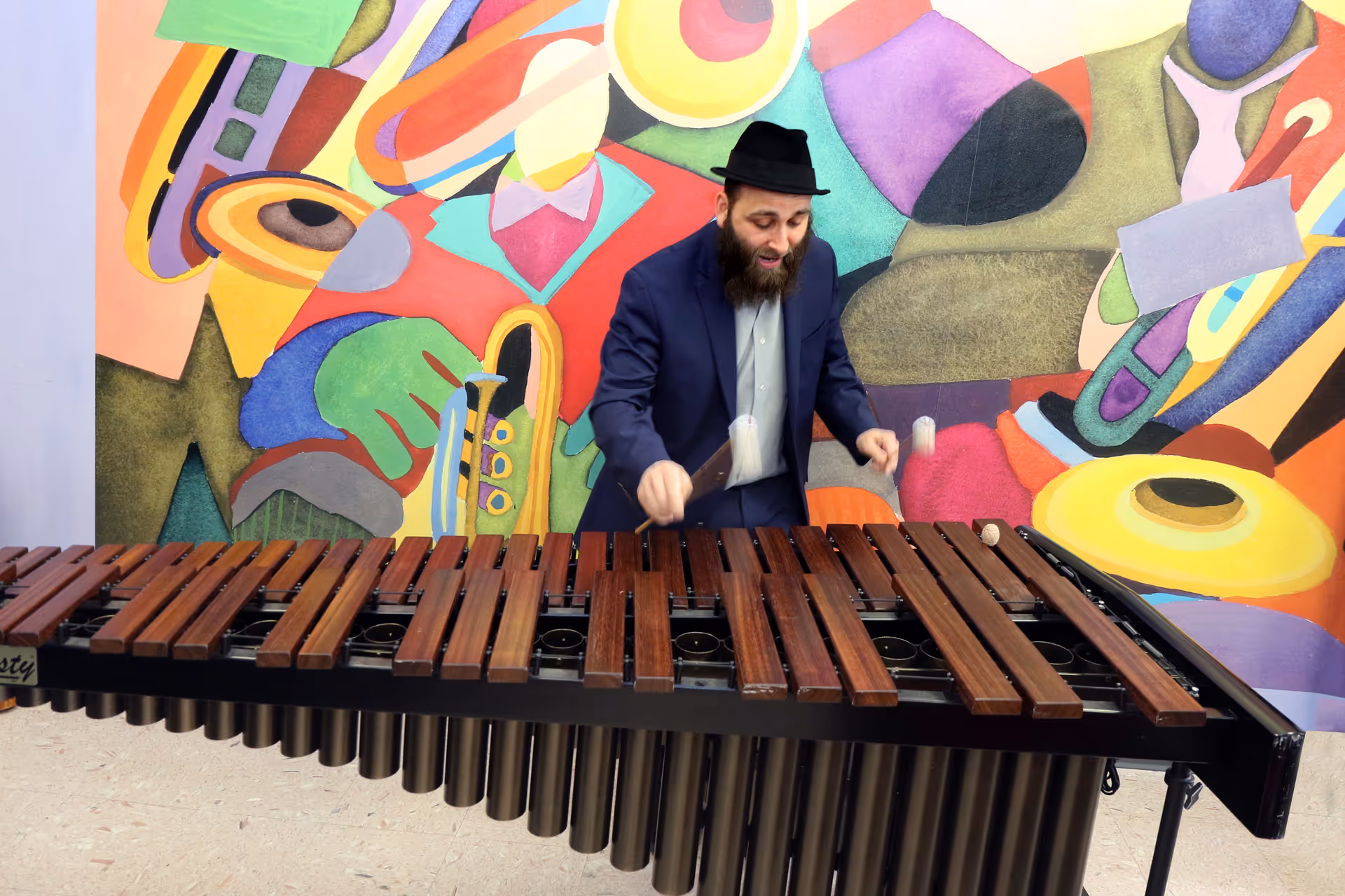A man wearing a dark suit and black hat is playing a marimba in front of a colorful mural featuring abstract shapes and musical instruments.
