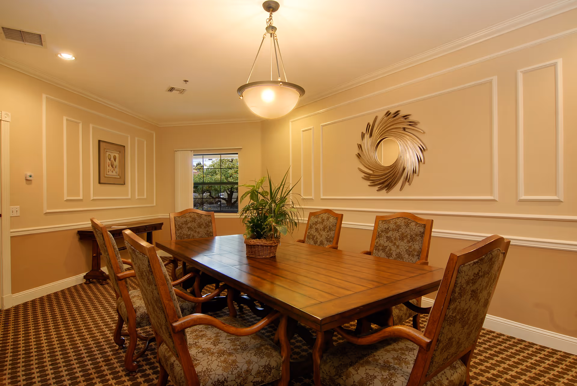 A warmly lit dining room with a wooden rectangular table surrounded by six upholstered chairs. A potted plant centerpiece sits on the table. The walls are beige with white trim and decorative molding, featuring a circular metallic wall art piece and a framed picture. A window with a view of greenery is visible in the background, and a hanging light fixture illuminates the room.