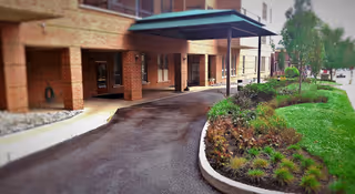 Entrance area of a building with a covered driveway and a landscaped garden bed with green plants and shrubs along the right side.