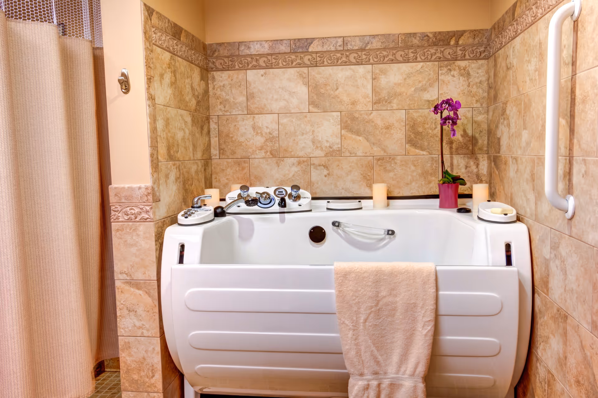 A walk-in bathtub with control knobs and a handlebar, surrounded by beige tiled walls. A beige towel hangs over the front of the tub, and there is a purple orchid plant and candles placed on the tub's edge. A grab bar is mounted on the right wall, and a shower curtain is partially visible on the left.