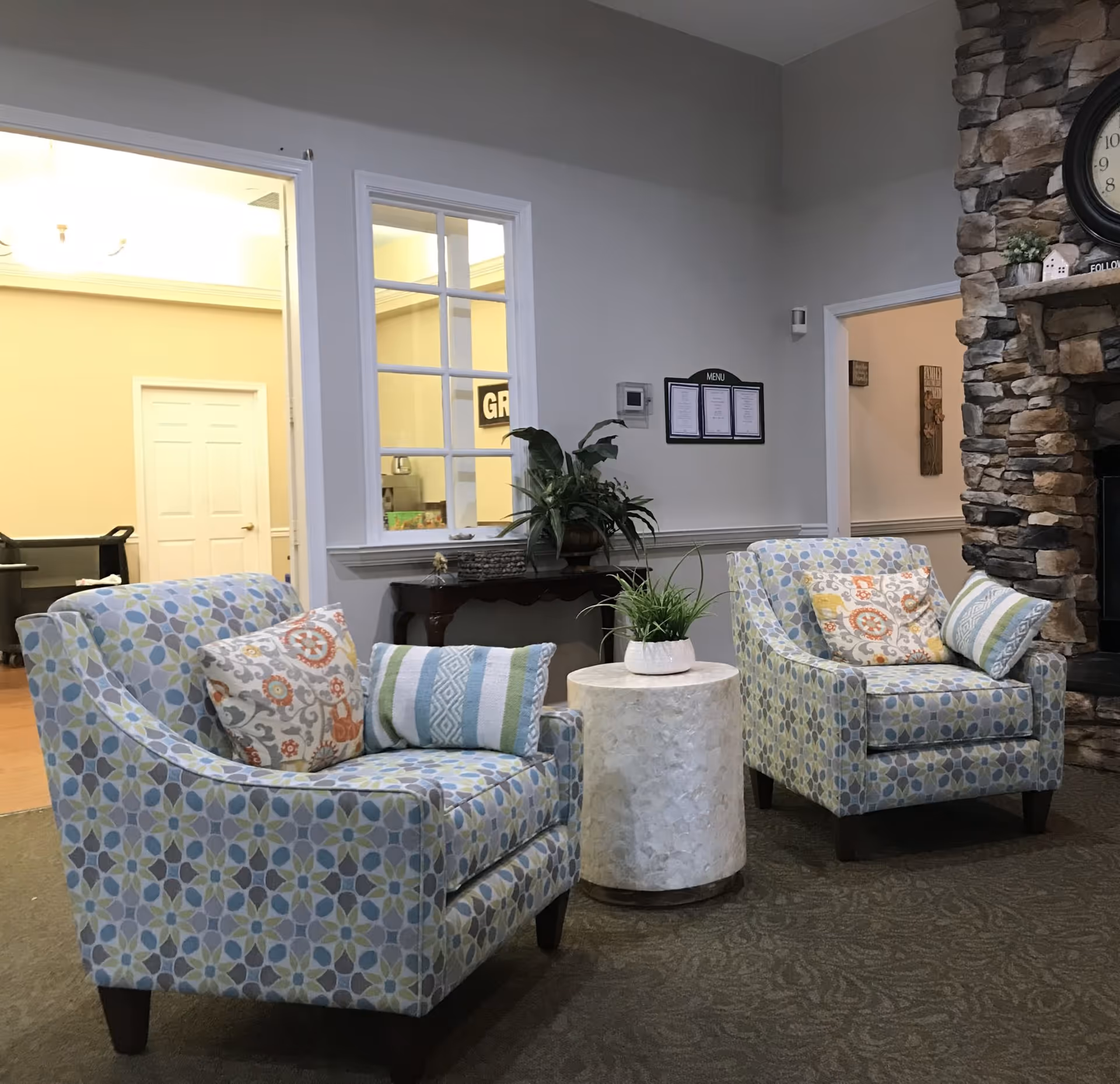 A cozy seating area in a senior living facility featuring two patterned armchairs with decorative pillows, a small round white table with a potted plant, a stone fireplace with a clock above it, and a wall-mounted menu. The room has neutral-colored walls and carpeted flooring.