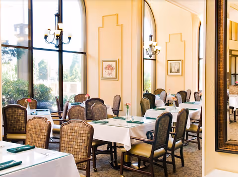 A bright dining room with multiple tables covered in white tablecloths, each set with green napkins and small flower vases. The room features large windows letting in natural light, elegant chandeliers, framed artwork on the walls, and a large mirror reflecting the space.