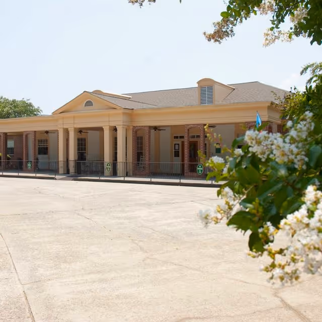 Front entrance of a single-story brick-and-column senior living facility with a covered portico and flowering shrubs in the foreground.