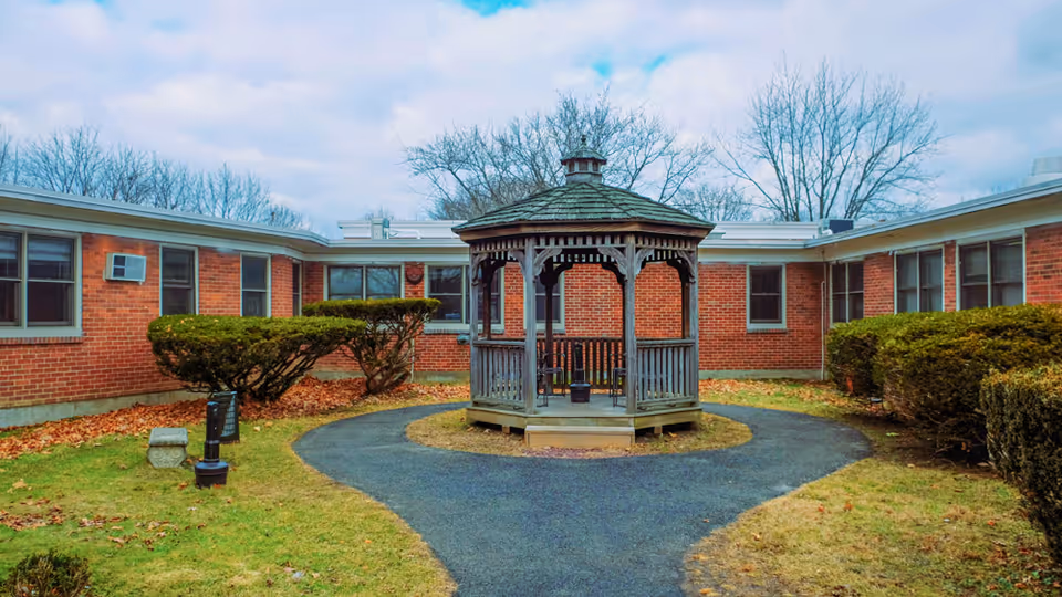 Wooden gazebo centered on a circular paved path in a brick single-story courtyard with trimmed shrubs and overcast sky.
