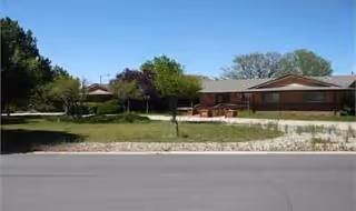 Single-story brick building with a front lawn, trees, and a paved street in the foreground.
