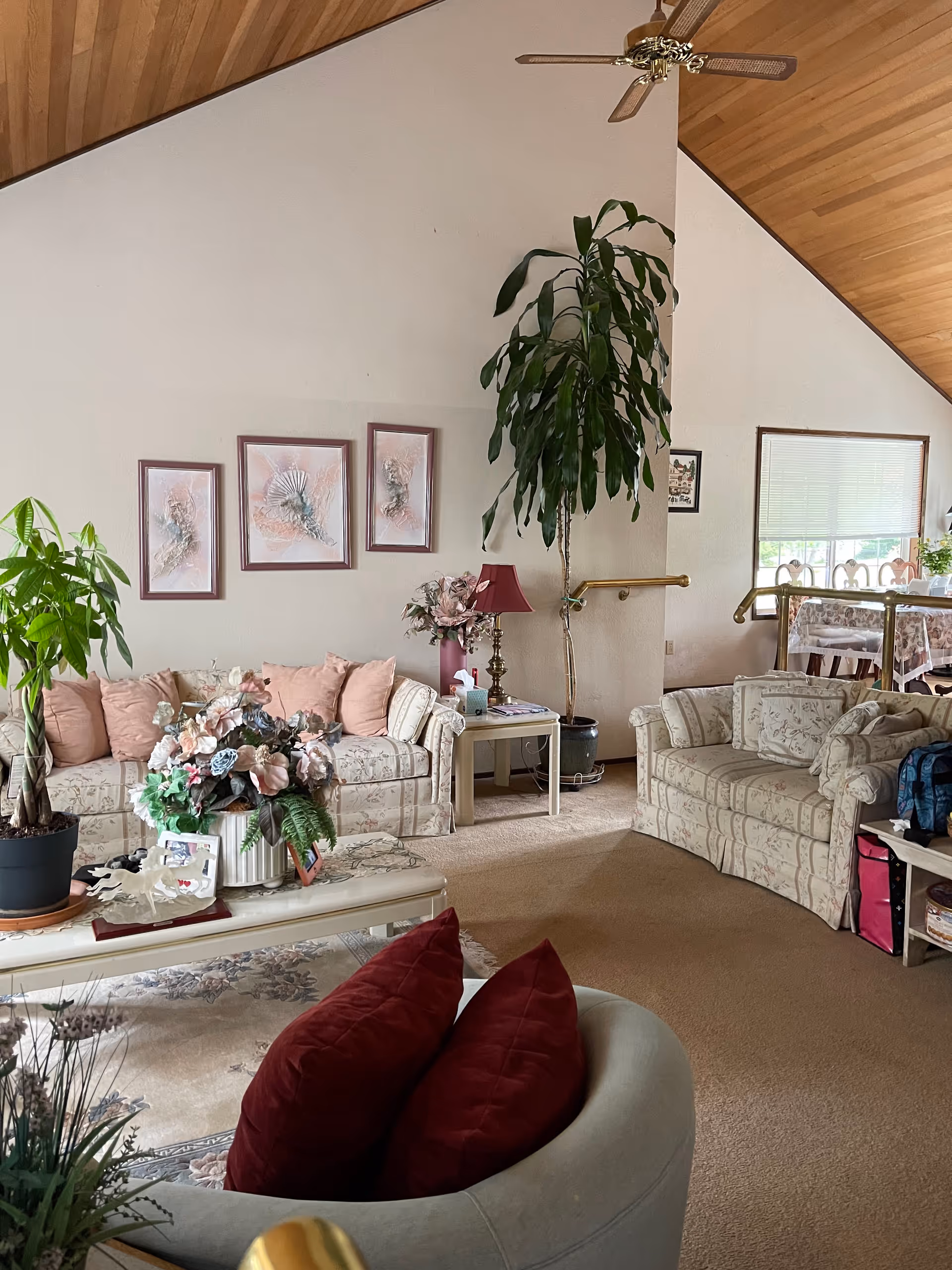 Spacious living room with floral-patterned sofas, a coffee table with floral arrangements, potted plants, and a vaulted wood ceiling with a ceiling fan.