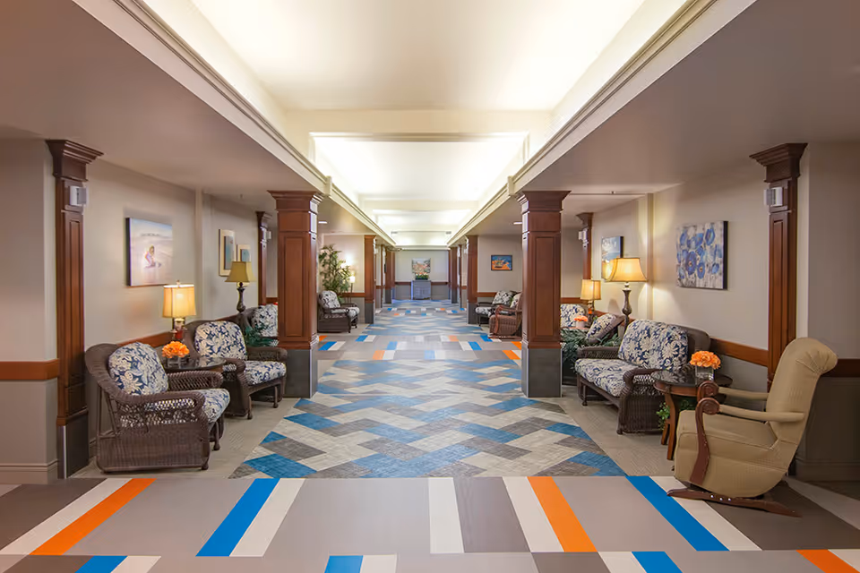 Long, well-lit interior hallway of a senior living facility with a patterned floor, wooden columns, and seating areas with chairs and lamps along both sides.