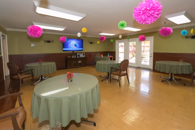 A common room with round tables covered in green tablecloths, each with a small flower arrangement. Several chairs are placed around the tables. Colorful paper decorations hang from the ceiling. A flat-screen TV is mounted on the far wall above a wooden cabinet. Double glass doors lead outside, allowing natural light into the room.