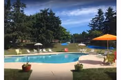 Outdoor swimming pool area with lounge chairs and umbrellas around the pool. There are trees and greenery in the background under a partly cloudy sky.