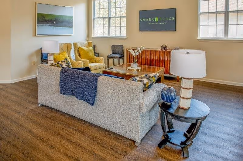 A cozy senior living room with a beige sofa adorned with patterned pillows and a blue throw blanket, two yellow armchairs with green cushions, a wooden coffee table with a vase of flowers, a side table with a lamp and decorative item, large windows letting in natural light, a wall-mounted TV displaying 'Amara Place', and a framed picture on the wall.