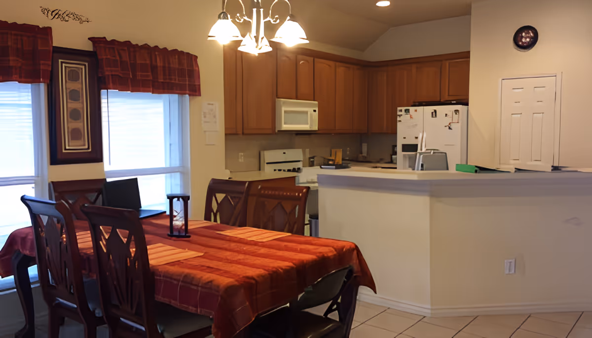 Interior view of a dining area and kitchen in a residential care facility. The dining area has a wooden table covered with a red and orange checkered tablecloth and six wooden chairs. The kitchen features wooden cabinets, a white microwave, a white stove, and a white refrigerator with magnets. There are two windows with red valances letting in natural light, and a chandelier with five lights hanging above the dining table.