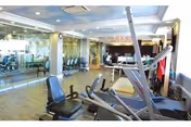 Interior view of a fitness room with exercise equipment including stationary bikes, elliptical machines, and weight machines. The room has large mirrors on one wall and windows on another, allowing natural light to enter. The floor is wooden, and the ceiling has recessed lighting.