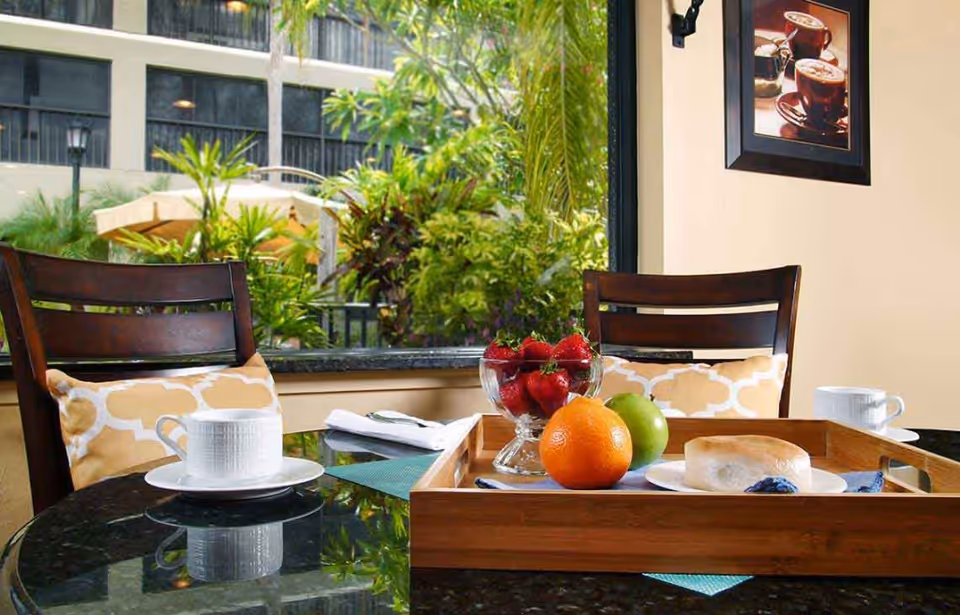 A cozy indoor dining area with a glass table set with a wooden tray holding a bowl of strawberries, an orange, a green apple, and a bagel on a plate. Two wooden chairs with yellow and white patterned cushions are positioned around the table. A large window behind the table shows lush green plants outside. A framed picture of coffee cups hangs on the beige wall.