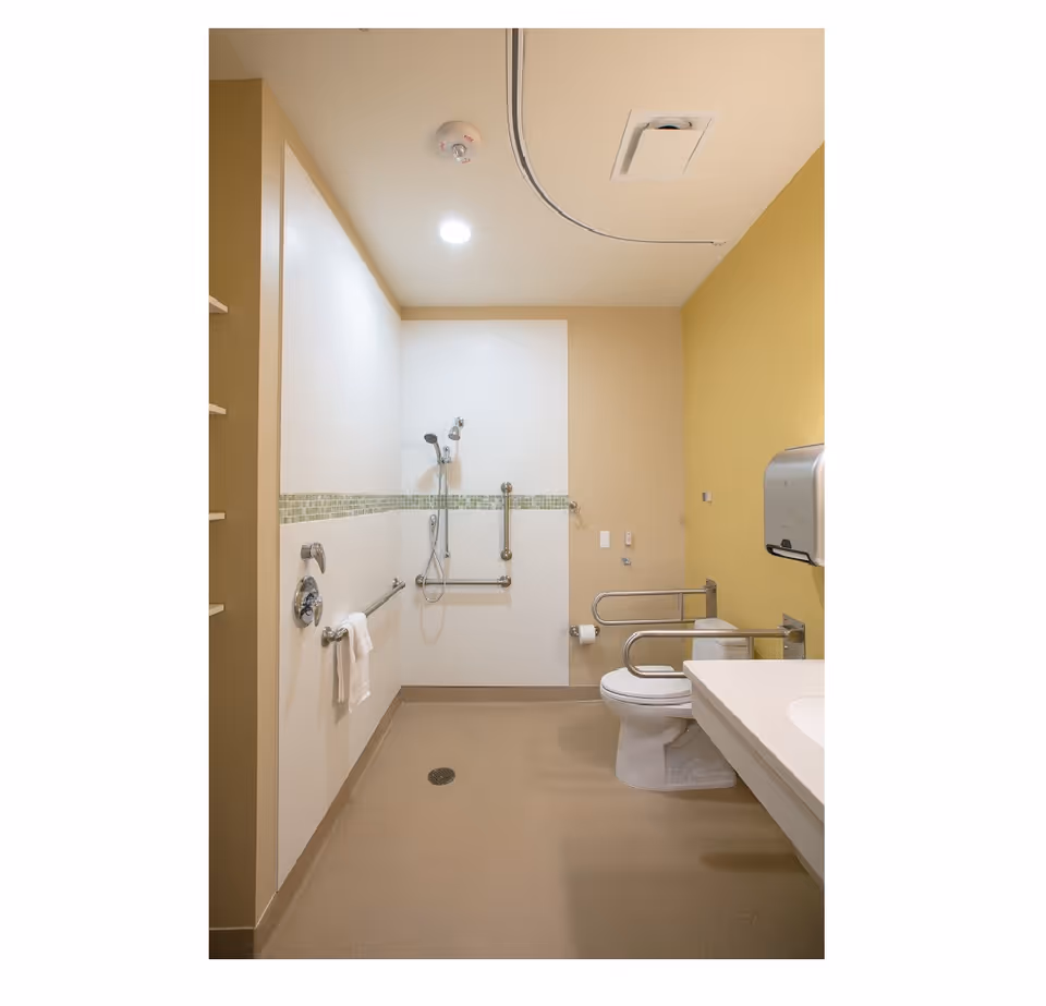 Accessible bathroom with a walk-in shower area featuring a handheld showerhead and grab bars. The room has a toilet with safety rails on both sides, a wall-mounted paper towel dispenser, and a countertop with a sink. The walls are painted in beige and yellow tones, and the floor is slip-resistant.
