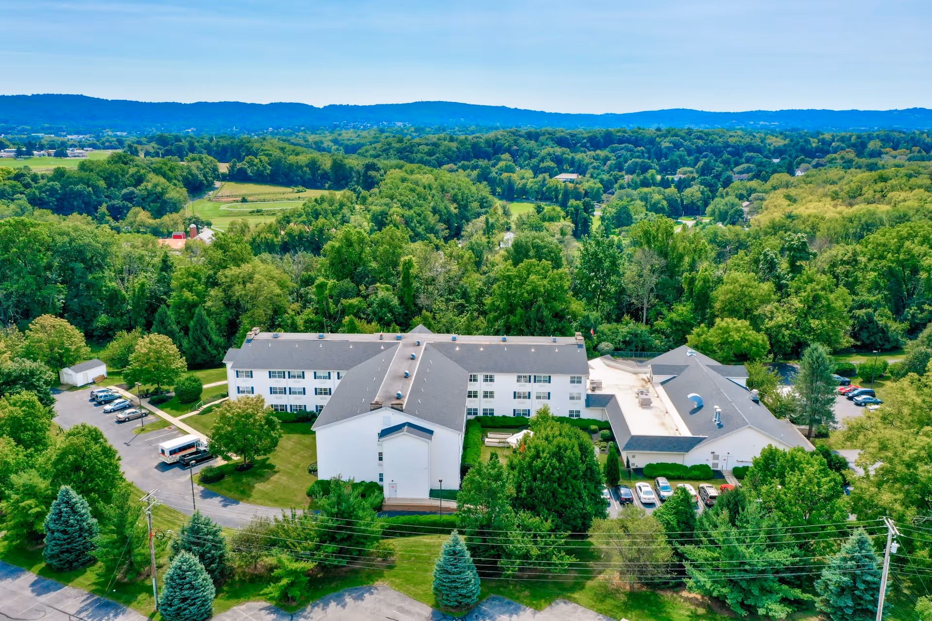 Aerial view of a senior living facility named Rittenhouse Village At Lehigh Valley surrounded by lush green trees and landscaping, with a parking area and a backdrop of rolling hills under a clear blue sky.