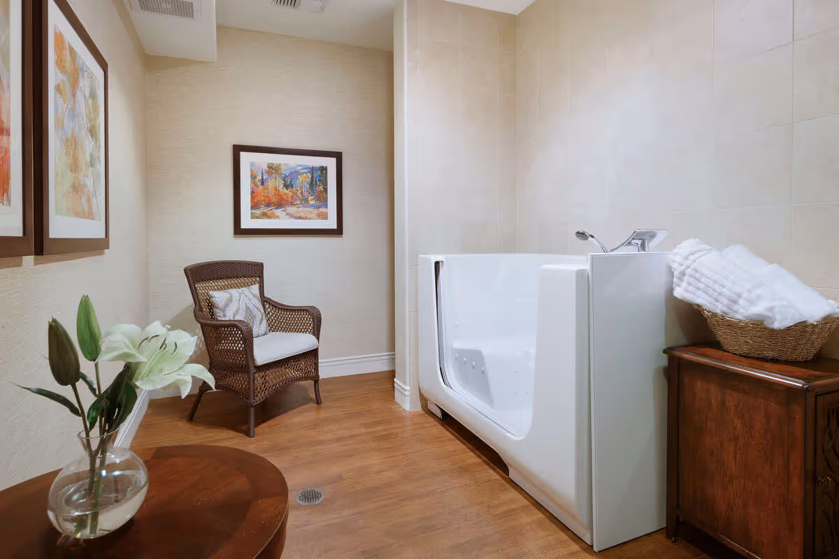 A bathroom with a walk-in bathtub on the right side, a wicker chair with a cushion against the back wall, framed artwork on the walls, a wooden cabinet with a basket of white towels, and a round wooden table with a glass vase holding white lilies.