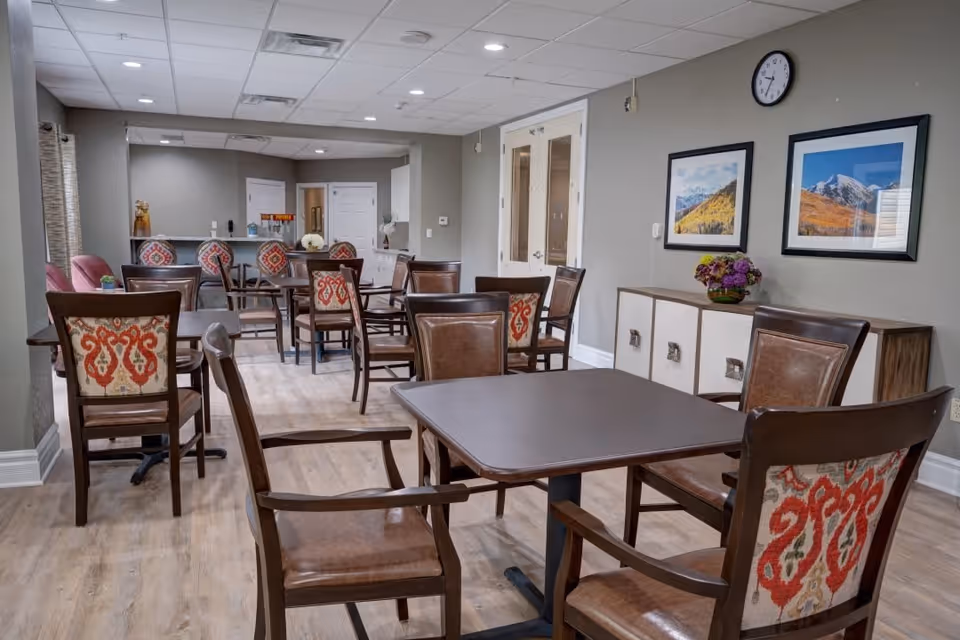 A dining room in a senior living facility with multiple wooden tables and chairs. The chairs have patterned upholstery on the backrests. The room has light wood flooring, gray walls, and two framed landscape pictures on the wall. A clock is mounted above the pictures, and a sideboard with decorative items is against the wall. The room is well-lit with ceiling lights.