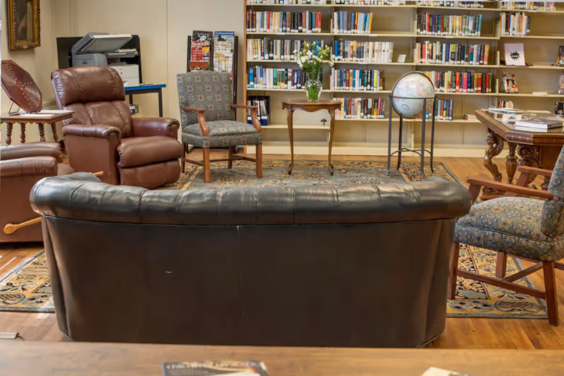 A cozy library or common area with a black leather couch in the foreground, two patterned armchairs, a brown leather recliner, a wooden table with a vase of flowers, a globe on a stand, and bookshelves filled with books in the background.