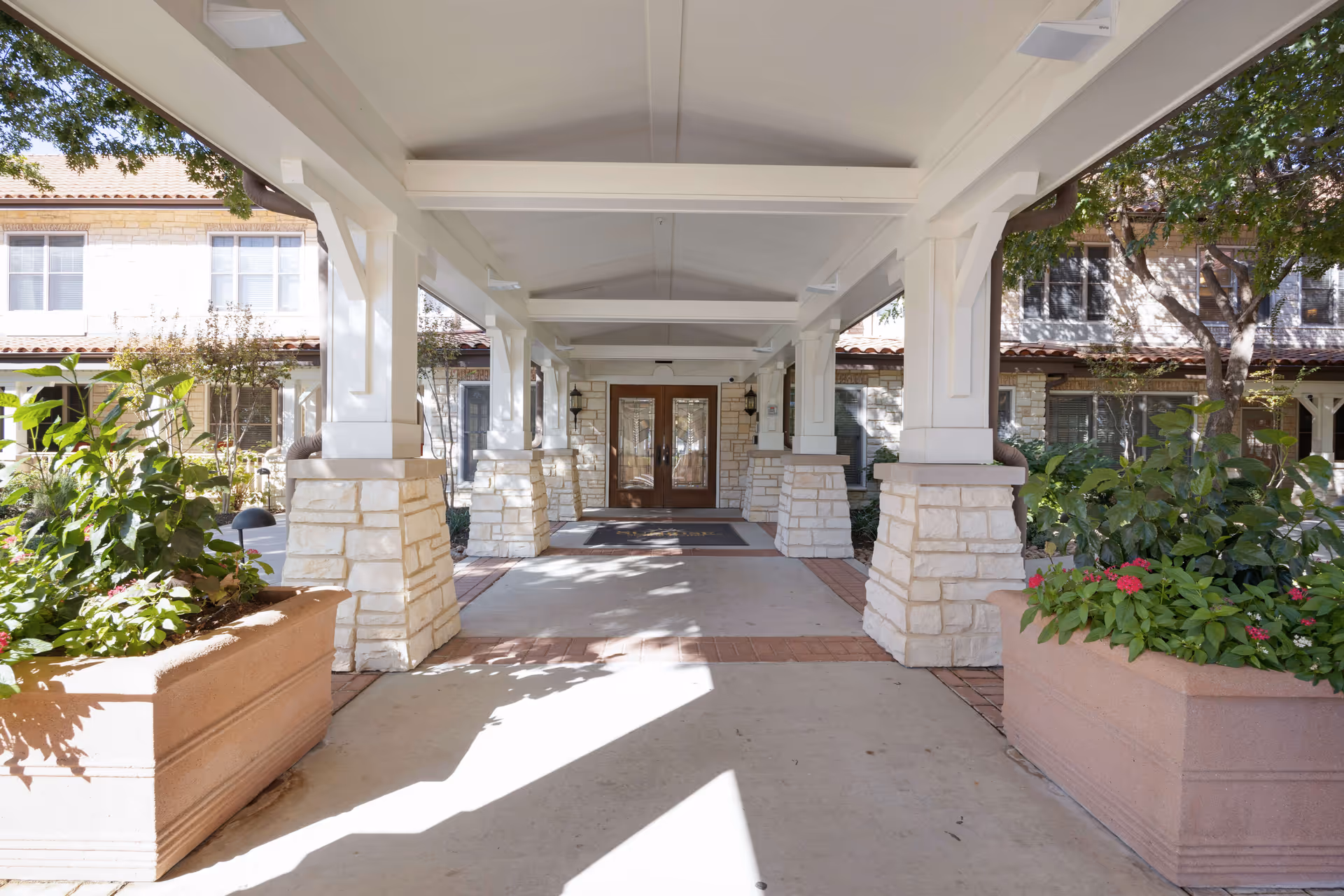 Covered walkway with white pillars and stone bases leading to double wooden doors of a building, with large planters filled with green plants and flowers on either side, and trees providing shade.