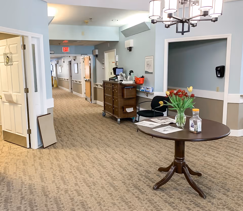 Interior hallway of a senior living facility with light blue walls and beige carpet. A round wooden table with a vase of red and yellow tulips, papers, and a bottle of juice is in the foreground. A dark wooden cart with office supplies and a computer monitor is against the wall. There are open doorways and a long corridor with handrails and wall lights.