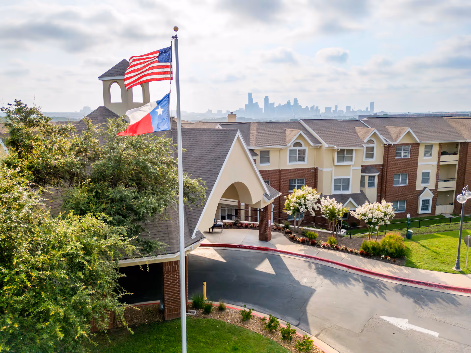 Exterior view of Brookdale Westlake Hills senior living facility with American and Texas flags flying on a flagpole. The building has a covered entrance, brick and beige siding, and landscaped grounds with trees and flowering bushes. The city skyline is visible in the background under a partly cloudy sky.