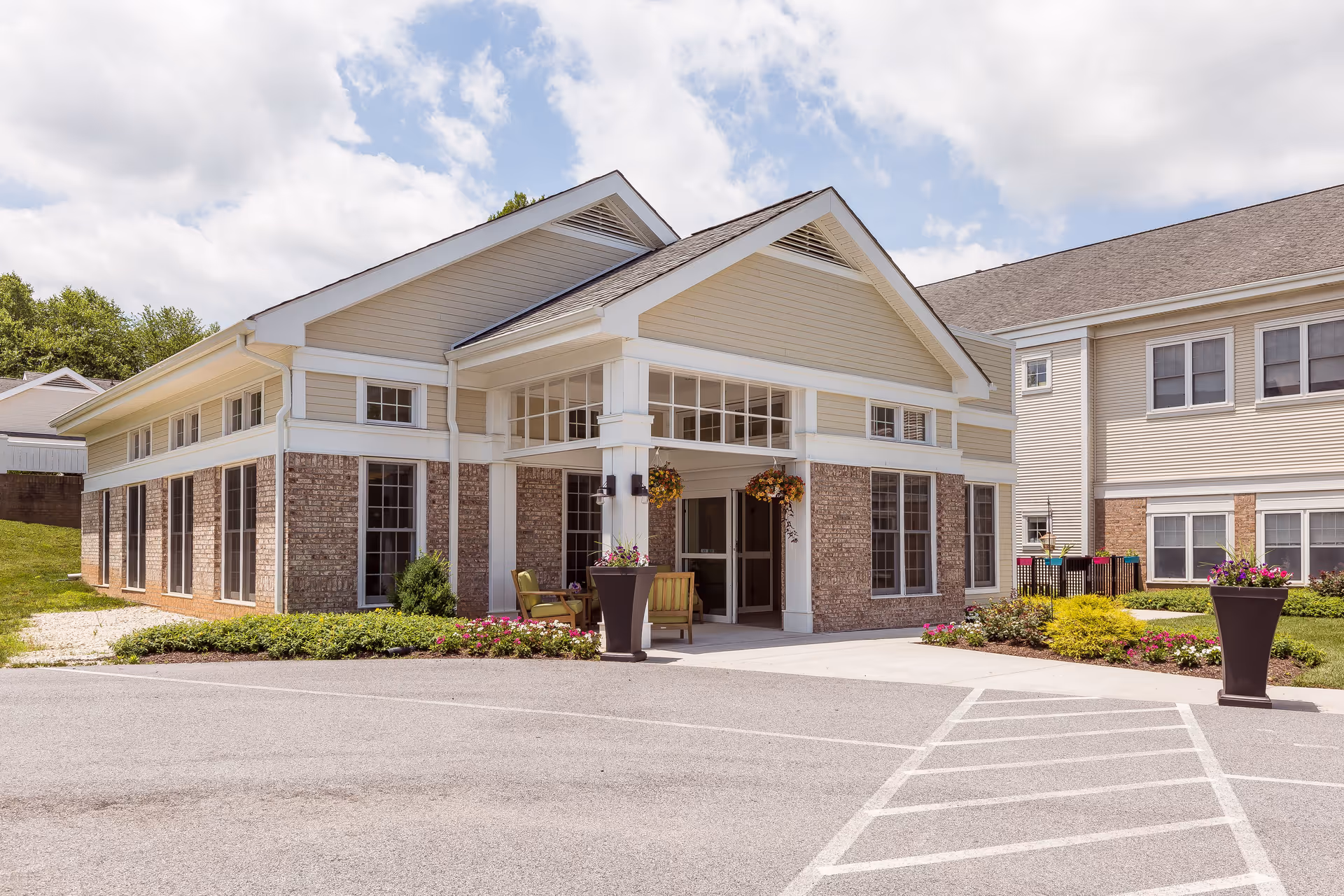 Exterior view of a senior living facility building with beige siding and brick accents. The entrance features a covered porch with hanging flower baskets and outdoor seating. There are landscaped flower beds and large planters with flowers near the entrance. The sky is partly cloudy.