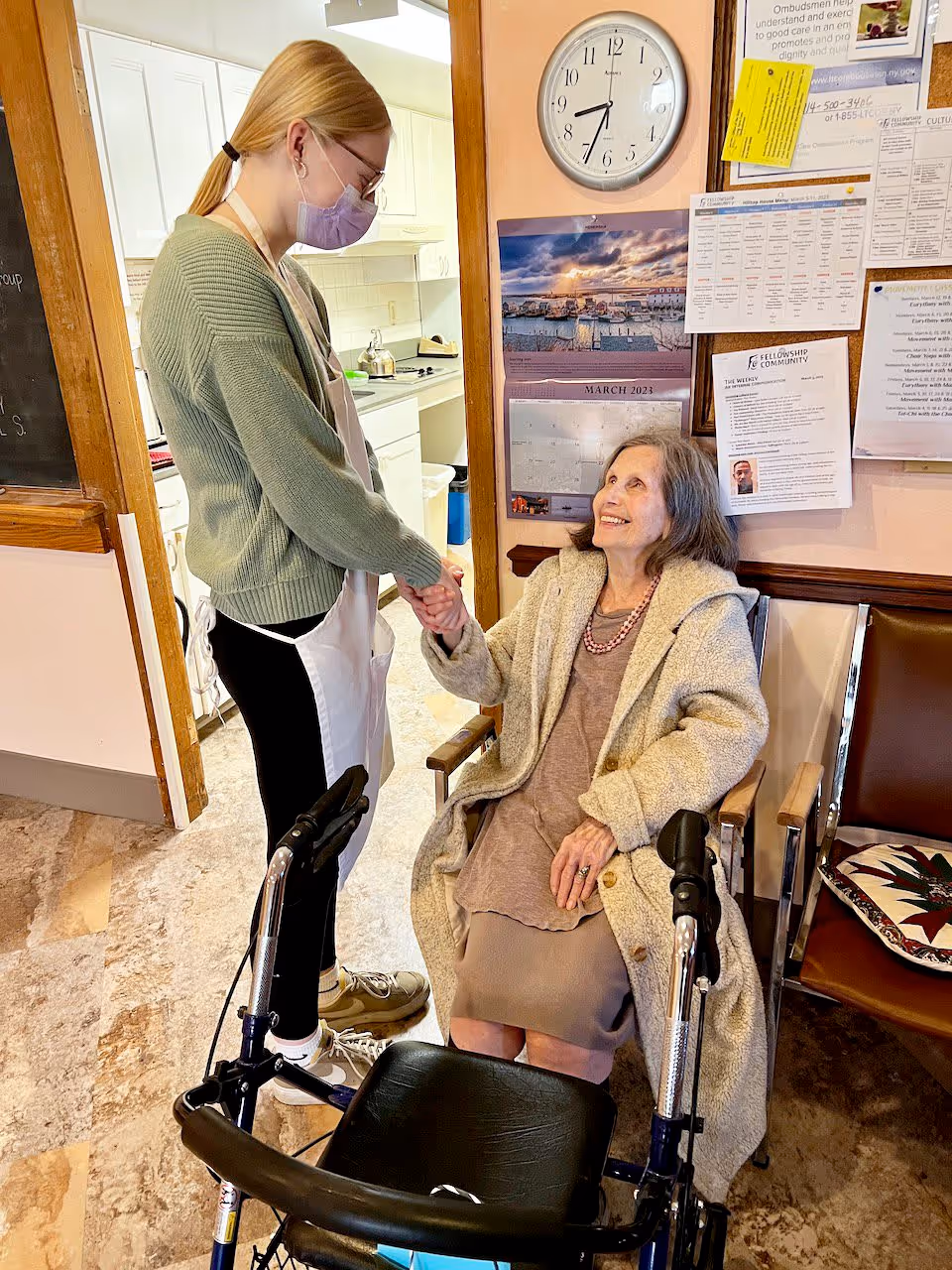 A young woman wearing a green sweater, apron, and face mask is holding hands and interacting warmly with an elderly woman seated in a wheelchair in an indoor setting. The elderly woman is wearing a beige coat and smiling up at the young woman. Behind them is a bulletin board with various notices and a clock showing the time as 7:40.