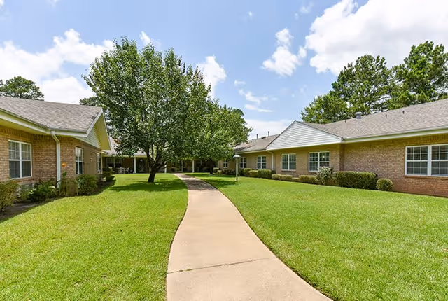 A paved walkway runs through a grassy courtyard with a large tree in the center, surrounded by single-story brick buildings under a partly cloudy blue sky.