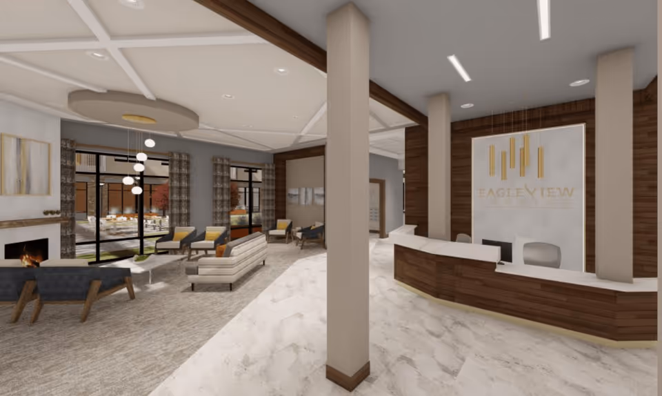 Interior view of a senior living facility lobby with a reception desk on the right featuring the Eagleview Landing logo on the wall behind it. The left side shows a seating area with modern chairs and sofas arranged around a coffee table and a fireplace, large windows with curtains reveal an outdoor patio area.