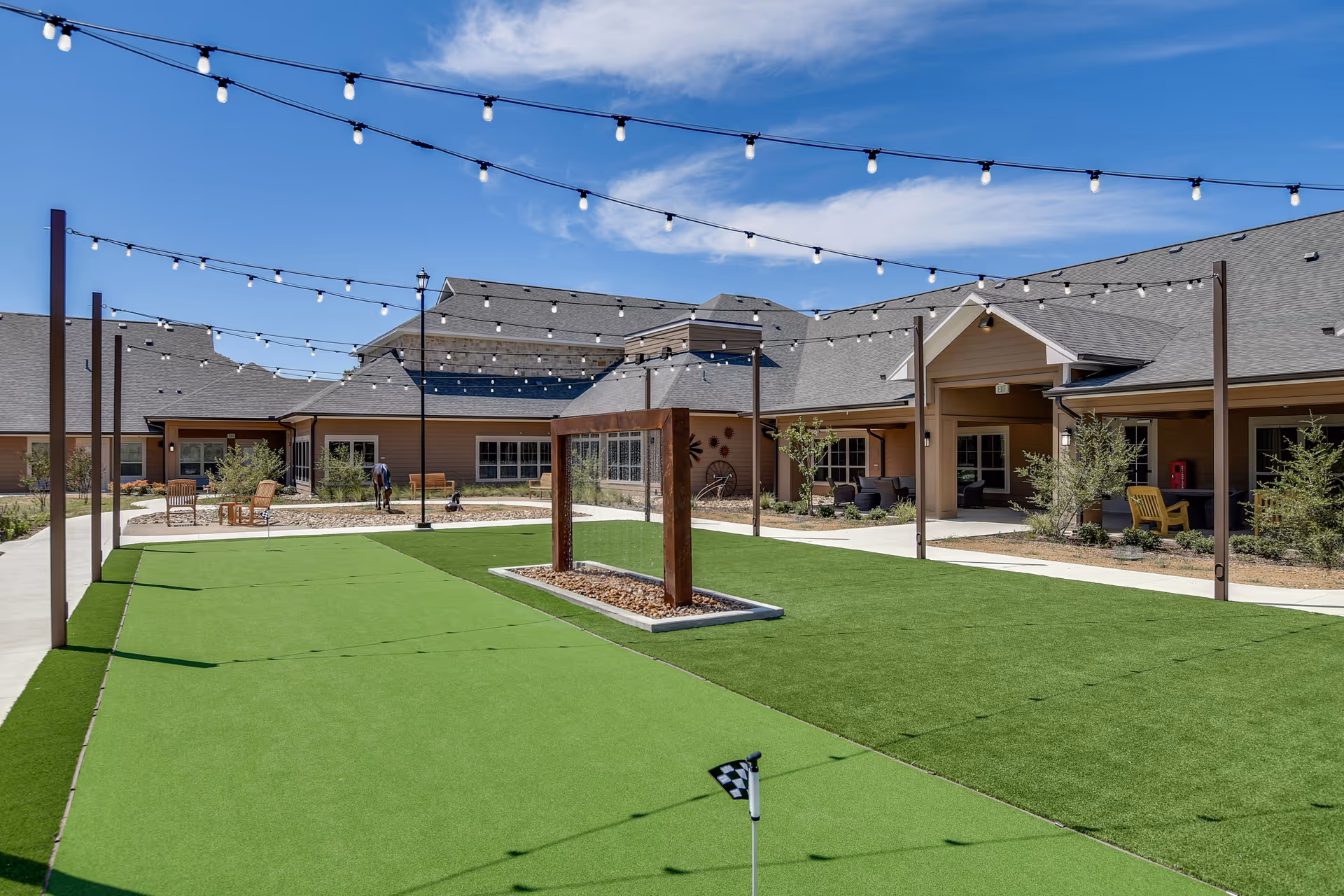 Outdoor courtyard area at Harvest of Roanoke with artificial green turf, string lights overhead, seating areas with chairs and tables, and a clear blue sky.