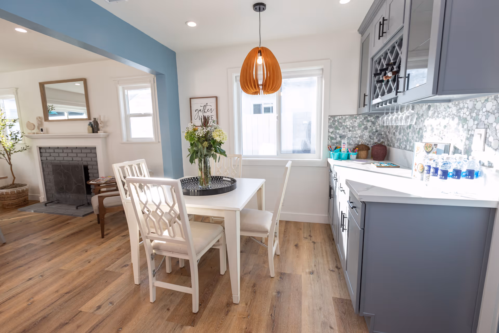 Bright dining area with a white table and chairs beside a gray kitchen and a wooden pendant light, with a fireplace visible in the adjacent living space.