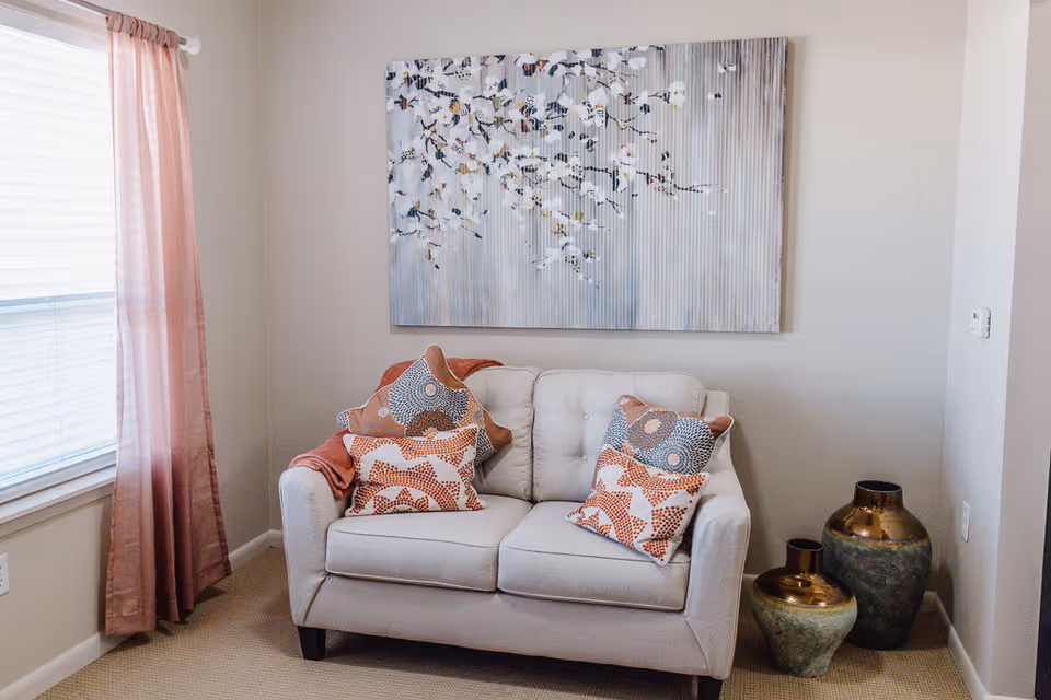 A small living room with a light loveseat topped with patterned pillows and a throw, floral wall art above, pink window curtains, and decorative vases on the floor.