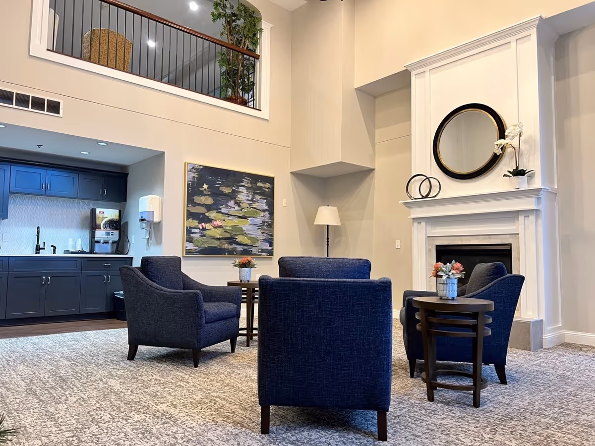 A cozy seating area in a senior living facility with four dark blue armchairs arranged around two small round tables with flower pots. Behind the seating area is a white fireplace with a round mirror and decorative items on the mantel. To the left, there is a kitchenette with dark blue cabinets, a coffee machine, and a wall-mounted paper towel dispenser. Above the kitchenette is a balcony with a railing and a large potted plant. The walls are painted light beige and there is a large painting of water lilies on the wall near the kitchenette.