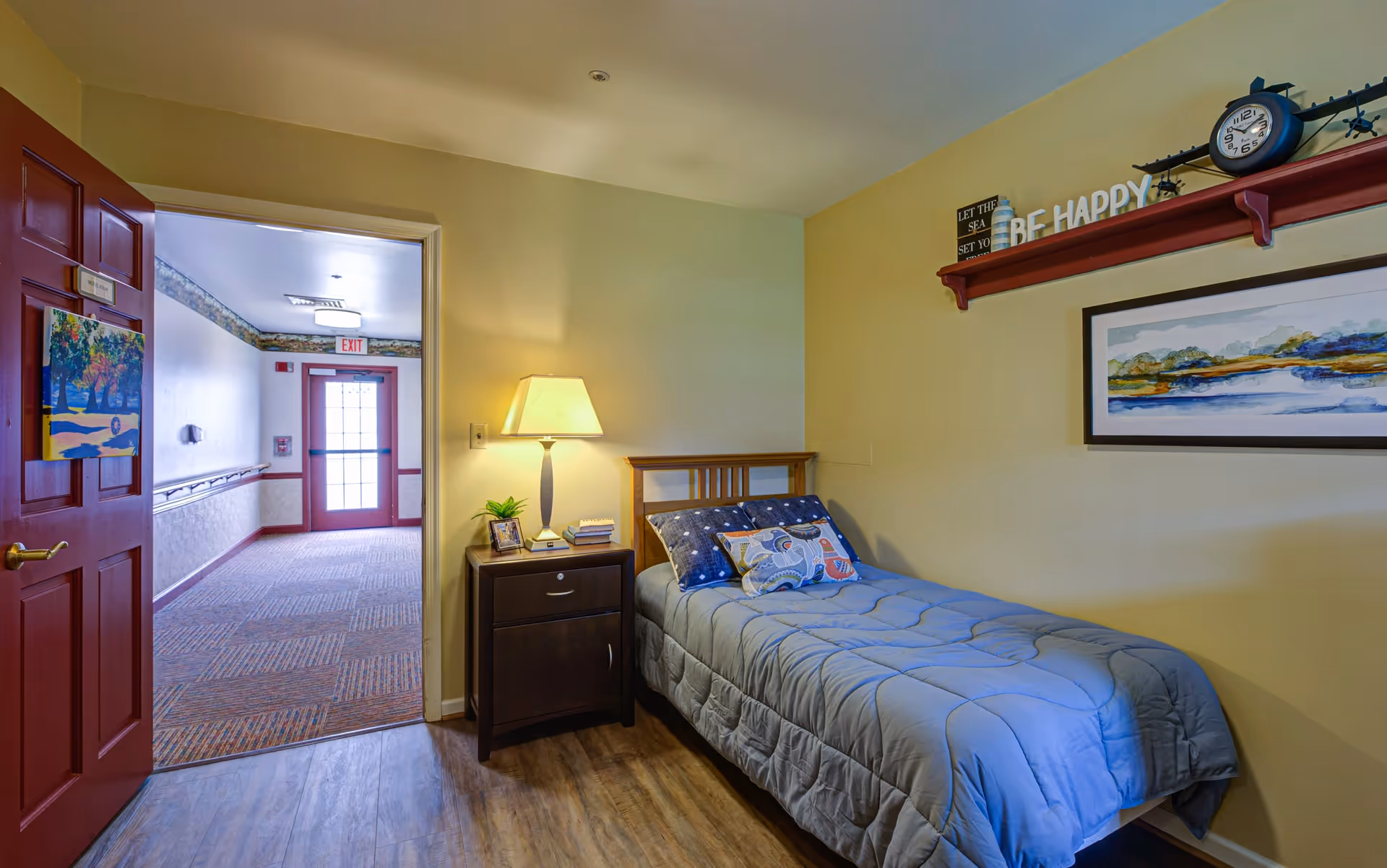 A small bedroom in a senior living facility with a single bed covered in a gray comforter and decorative pillows. Next to the bed is a dark wooden nightstand with a lamp, a small plant, and some books. The walls are painted light yellow and decorated with a framed landscape painting and a shelf holding a clock and decorative letters spelling 'BE HAPPY'. The door to the room is open, showing a hallway with carpeted flooring and an exit door at the end.