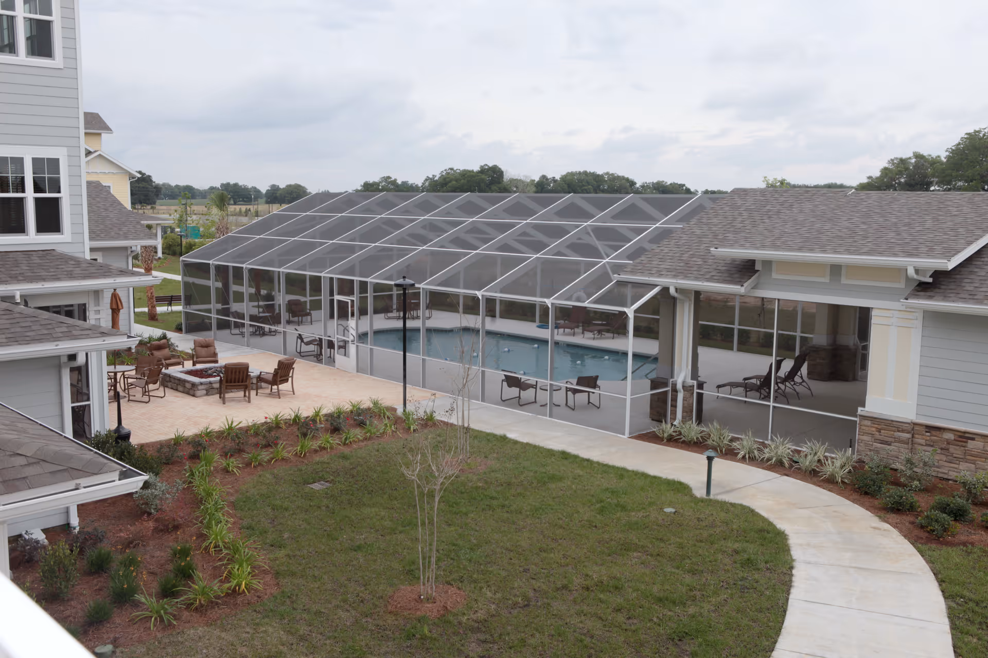 Outdoor view of a senior living facility featuring a screened-in swimming pool area with lounge chairs inside. Surrounding the pool enclosure is a patio with several chairs and a fire pit. The buildings have light gray siding with stone accents and multiple windows. There is a curved concrete walkway and landscaped greenery in the foreground under a cloudy sky.