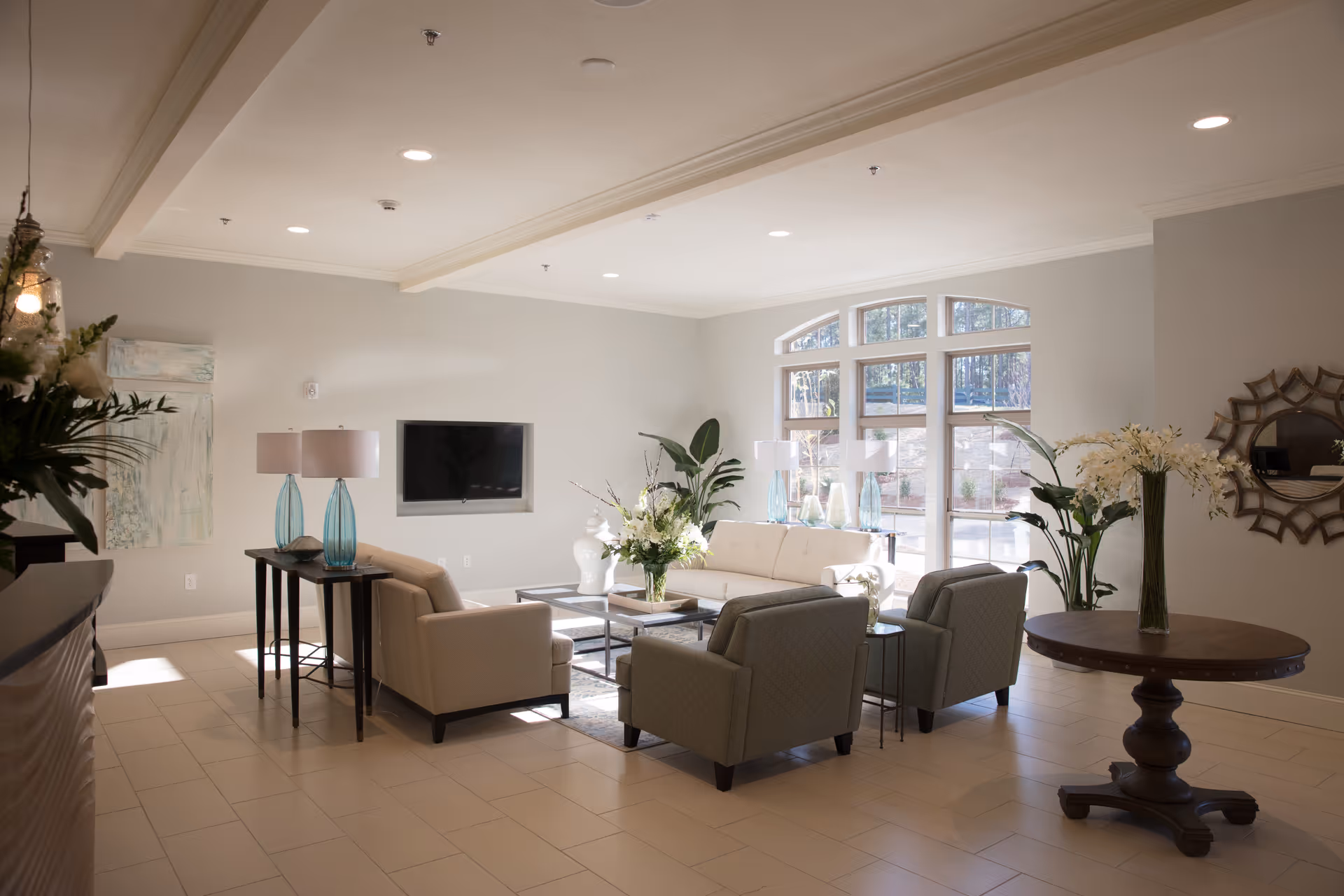 A bright and spacious living room area with large windows letting in natural light. The room features a white sofa, two armchairs, a coffee table with a floral arrangement, side tables with lamps, and decorative plants. There is a wall-mounted flat-screen TV and a round wooden table with a vase of flowers. The walls are painted light gray and the floor is tiled.
