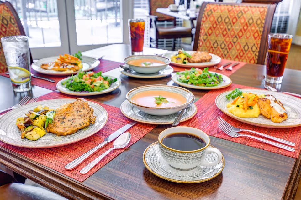 A dining table set with plates of food including grilled chicken, mixed vegetables, salads, bowls of soup, glasses of iced tea, and a cup of black coffee. The table has red placemats and is in a well-lit room with patterned chairs and glass doors in the background.