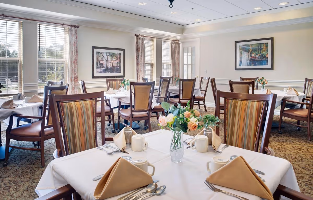 A bright dining room with several tables covered in white tablecloths, each set with beige napkins, silverware, mugs, and small floral centerpieces. The room has large windows with curtains, framed artwork on the walls, and patterned carpet flooring.