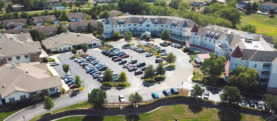 Aerial view of Victorian Village Continuing Care Senior Community showing multiple connected buildings with light-colored roofs and turrets, a large parking lot filled with cars, surrounding trees, and nearby residential houses.