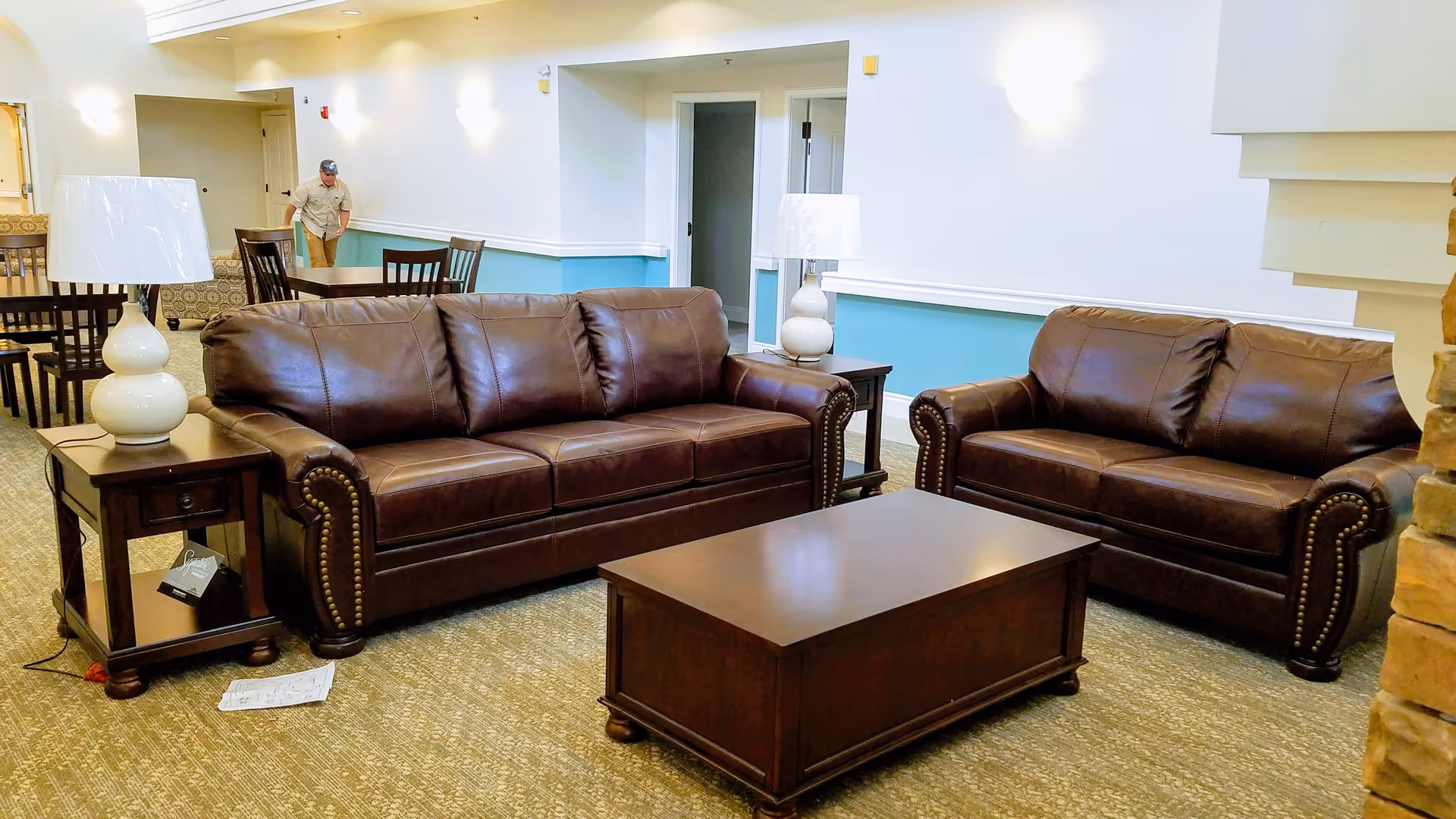 A seating area in a senior living facility featuring two brown leather sofas, a wooden coffee table, and two side tables with white lamps. In the background, there are dining tables and chairs, and a person walking down a hallway.