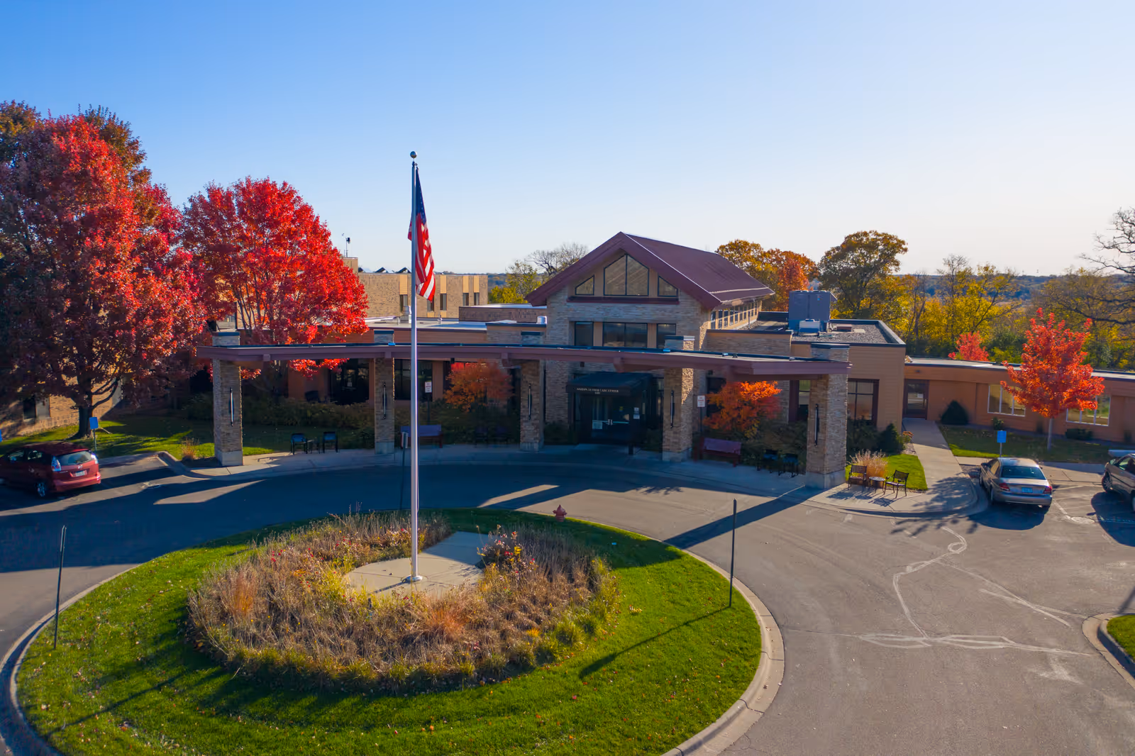Front entrance of a senior living facility with a circular driveway, central flagpole, and autumn trees.