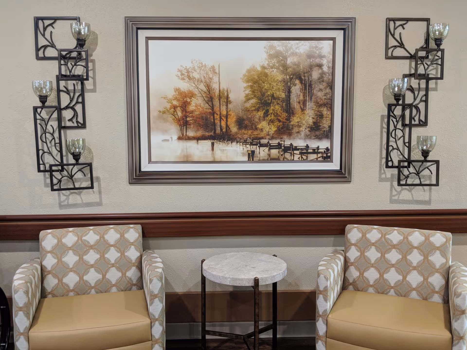 Two patterned armchairs with beige cushions are positioned on either side of a small round marble-top table against a beige wall. Above the table hangs a framed painting depicting a misty autumn scene with trees and a wooden dock extending into the water. On each side of the painting, there are decorative black metal wall sconces holding glass candle holders.