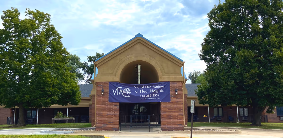Front exterior view of Via of Des Moines at Fleur Heights facility, showing a brick building with a peaked roof entrance, a banner with the facility name and contact information, and large green trees on either side.