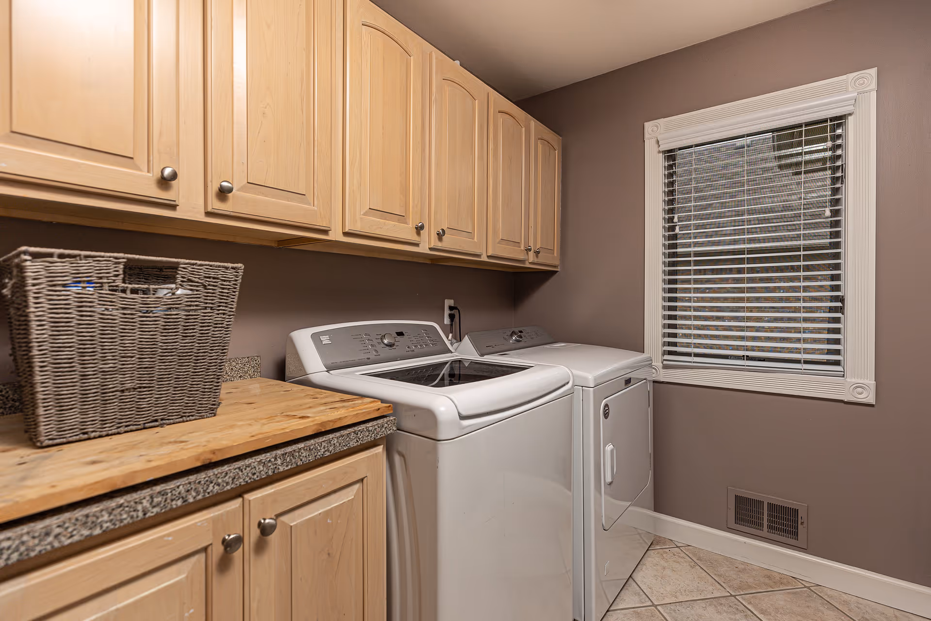 Laundry room with a washing machine and dryer side by side, wooden cabinets above and below a countertop, a wicker basket on the counter, and a window with blinds on the right wall.